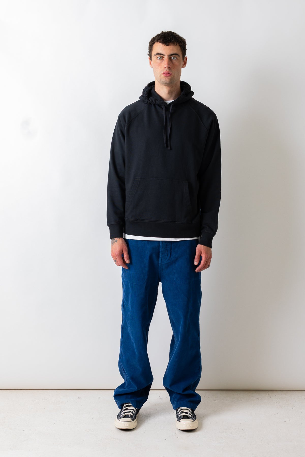 A man stands against a plain white studio wall wearing a black pullover hoodie, blue pants, and black-and-white sneakers.