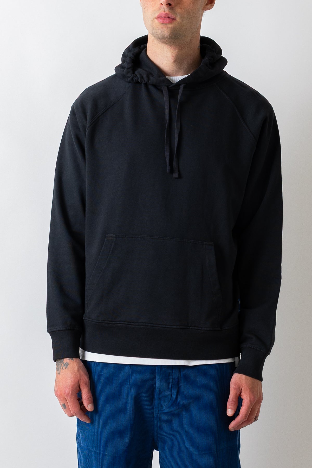 A man wearing a black pullover hoodie with a kangaroo pocket and drawstrings, layered over a white t-shirt and paired with blue jeans, photographed against a light gray background.