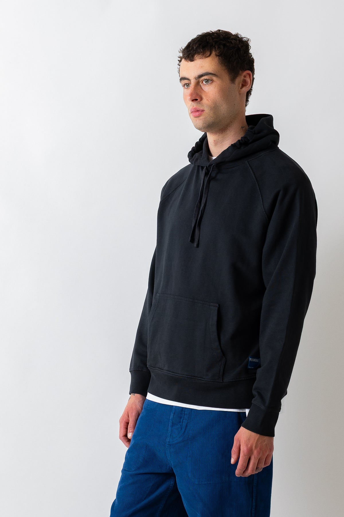 A man wearing a black pullover hoodie with a kangaroo pocket and drawstrings, paired with blue pants, standing against a light gray studio background.