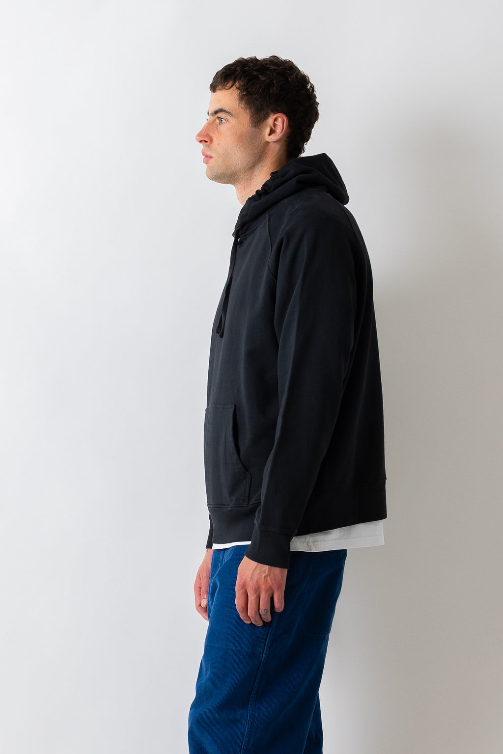 A man in profile wearing a dark navy pullover hoodie over a white t-shirt and blue jeans, standing against a plain light gray wall.