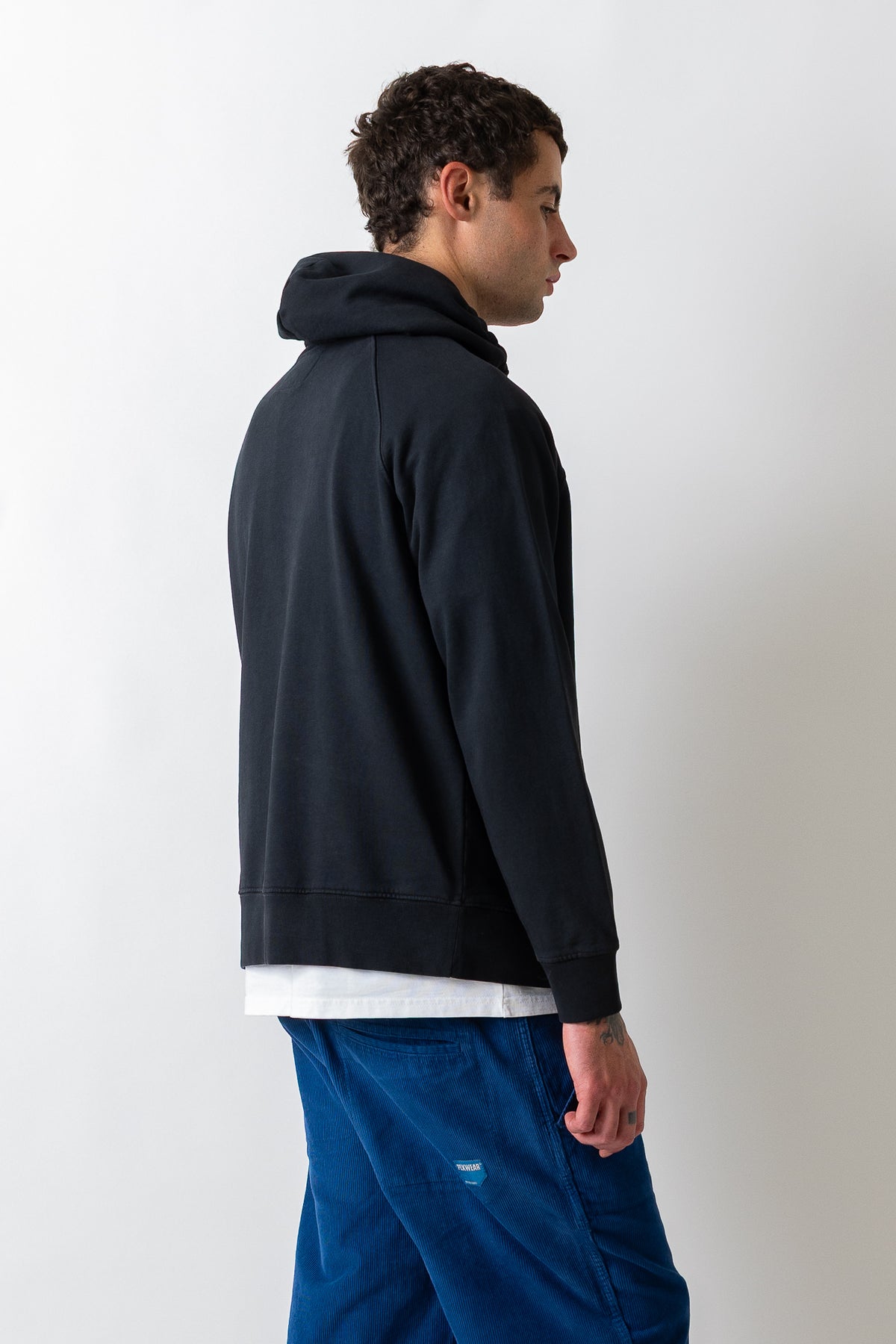 A man shown from the side profile wearing a dark navy pullover hoodie, a white t-shirt, and blue corduroy pants against a plain light gray background.