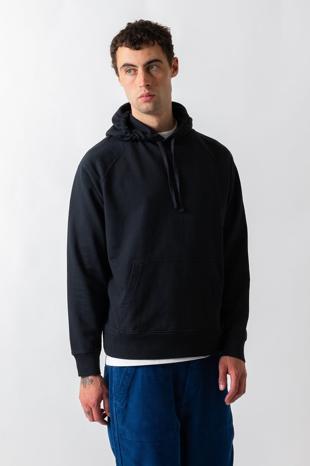 A man wearing a black pullover hoodie with a kangaroo pocket and drawstrings, paired with blue pants, standing against a light gray studio background.