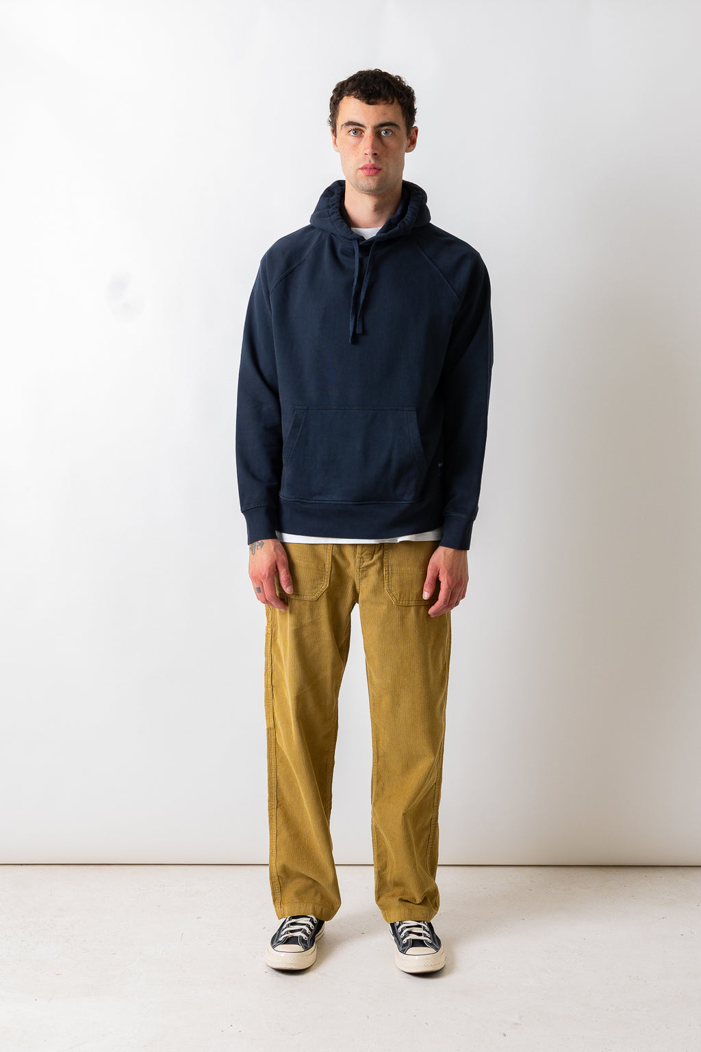 A man stands against a plain off-white studio wall wearing a dark navy pullover hoodie, mustard yellow corduroy pants, and black-and-white sneakers.