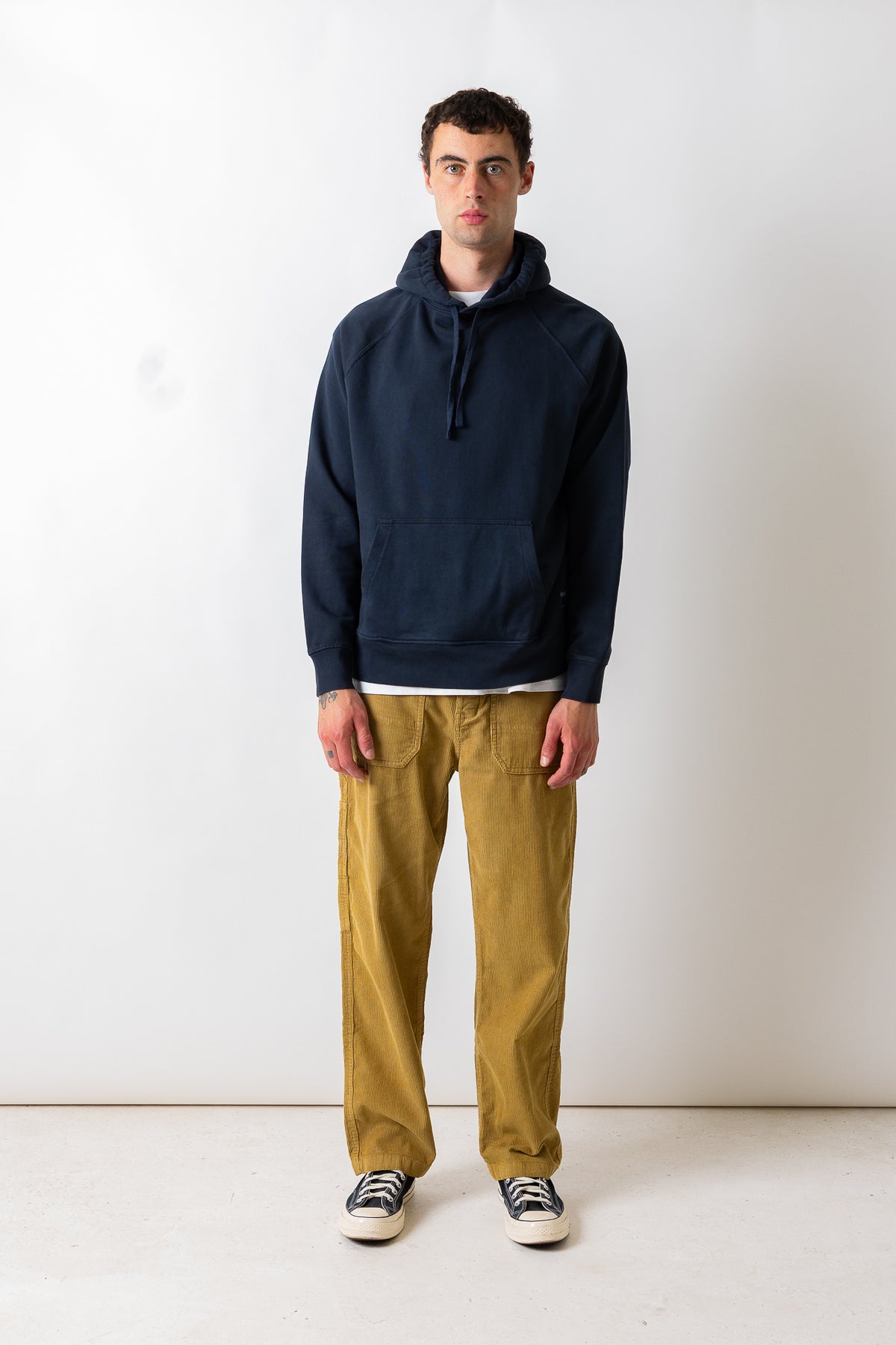 A man stands against a plain off-white studio wall wearing a dark navy pullover hoodie, mustard yellow corduroy pants, and black-and-white sneakers.