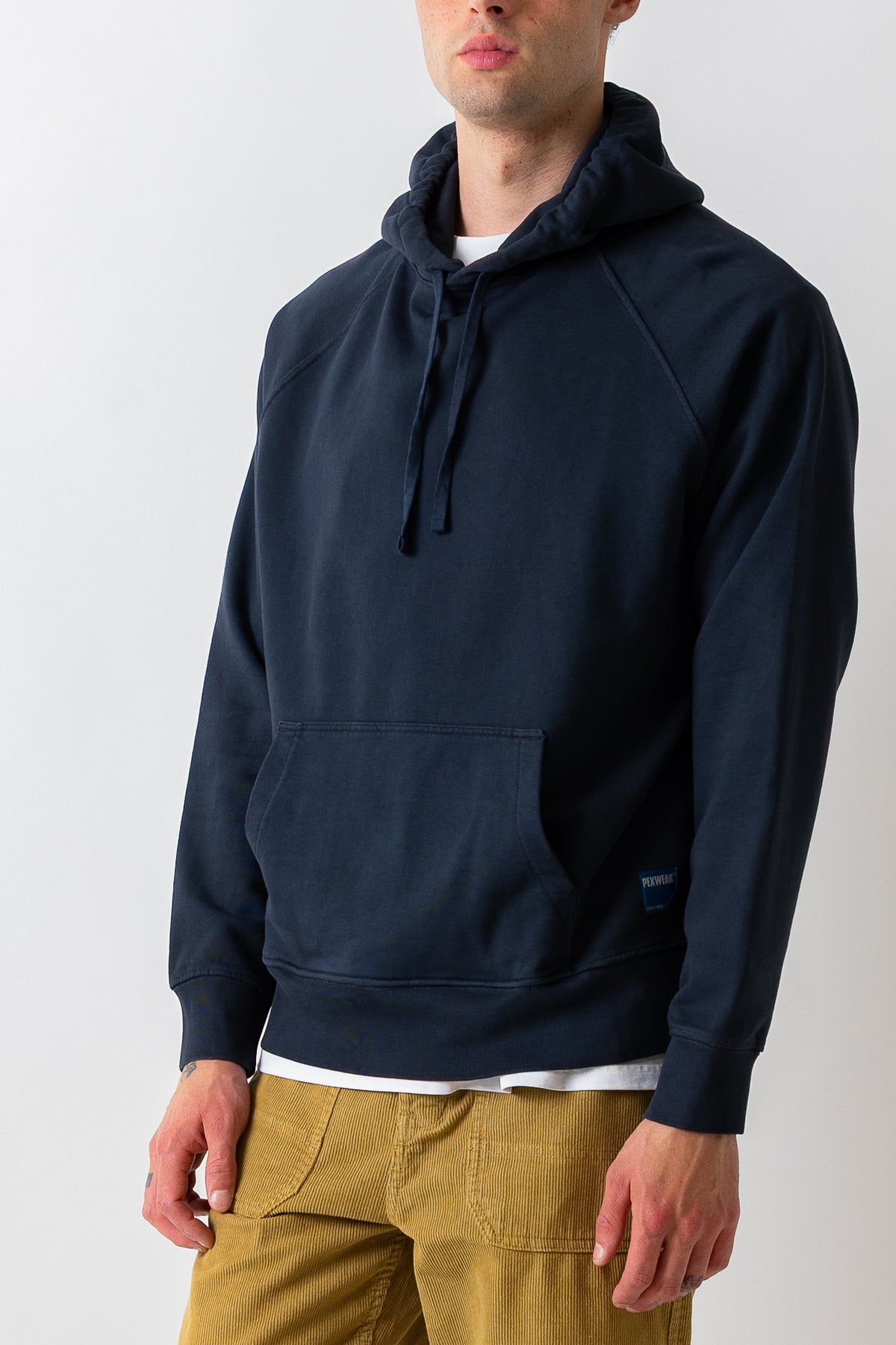 A man wearing a navy blue pullover hoodie with a kangaroo pocket and drawstrings, layered over a white t-shirt and paired with mustard yellow pants, photographed against a light gray studio background.