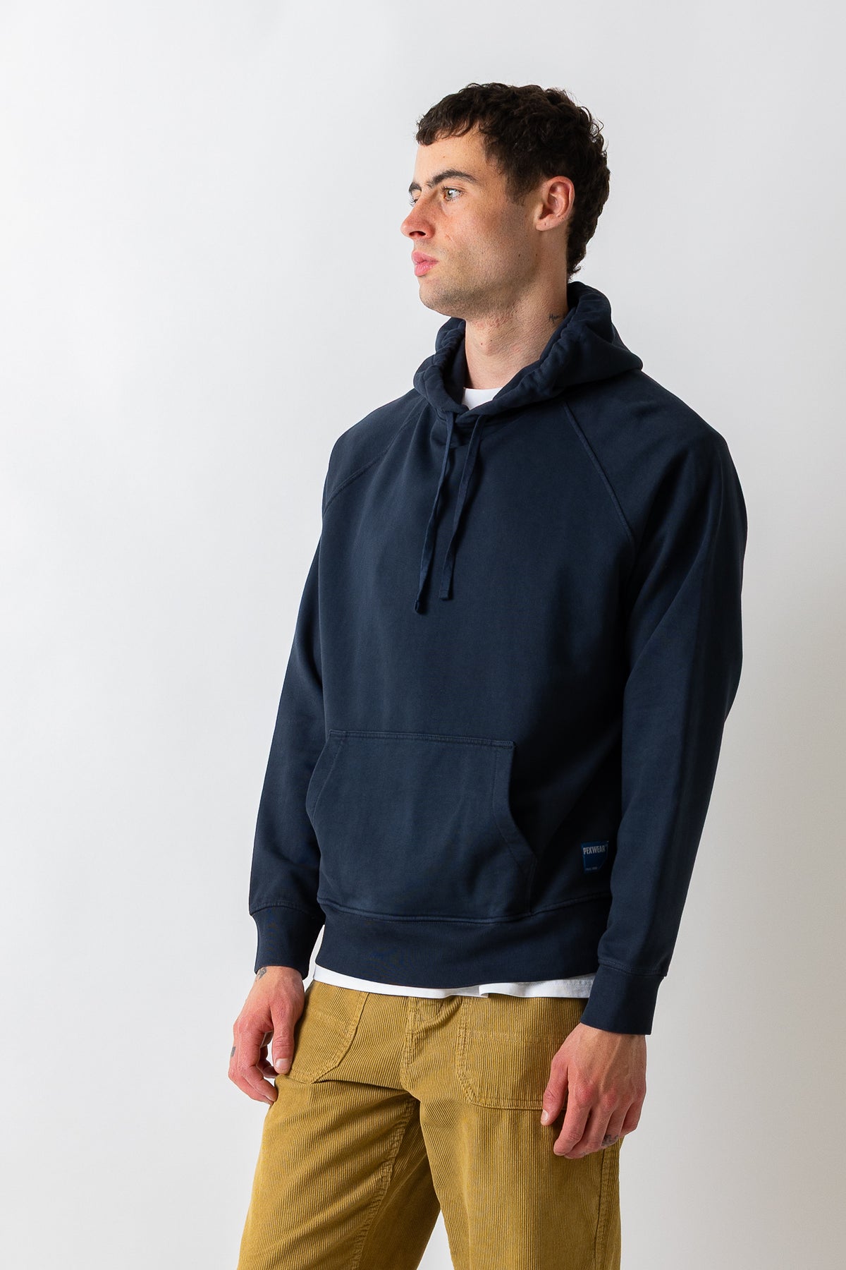 A man wearing a navy blue pullover hoodie with a kangaroo pocket and drawstrings, paired with mustard yellow pants, standing against a light gray studio background.