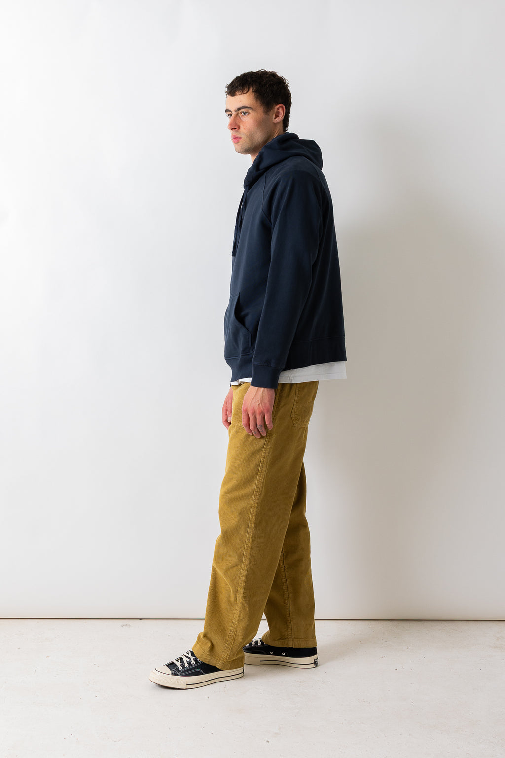 A man standing in profile wearing a navy blue hoodie, a white t-shirt, mustard yellow pants, and black canvas sneakers against a plain white studio background.
