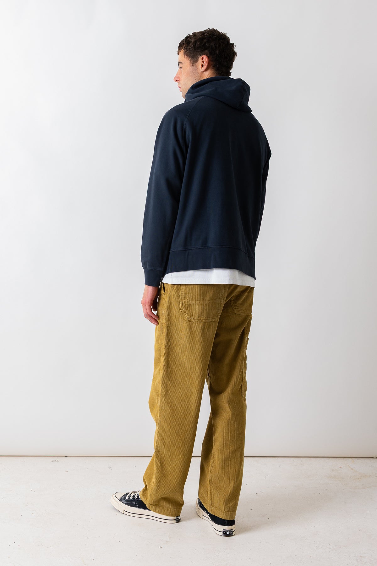 A man standing in profile wearing a dark navy hoodie, a white t-shirt, mustard yellow pants, and black canvas sneakers against a plain white studio background.
