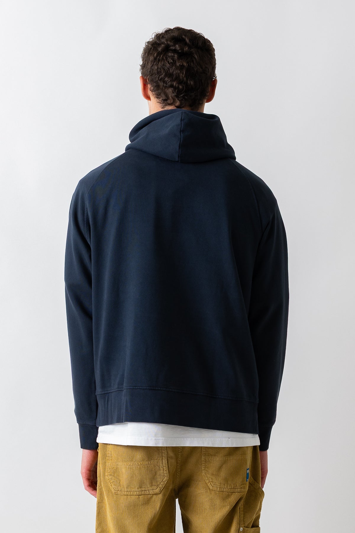 A man viewed from behind wearing a dark navy blue hooded sweatshirt, a white t-shirt, and khaki pants against a light gray studio background.