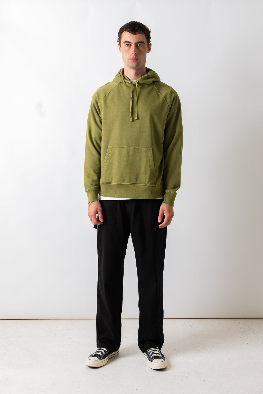A man wearing an olive green pullover hoodie with a front kangaroo pocket, paired with black pants and black-and-white sneakers, standing against a plain white studio background.