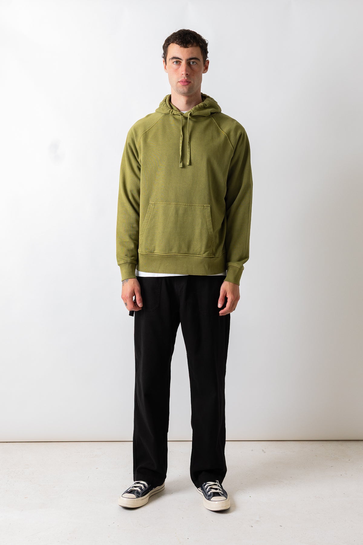 A man wearing an olive green pullover hoodie with a front kangaroo pocket, paired with black pants and black-and-white sneakers, standing against a plain white studio background.