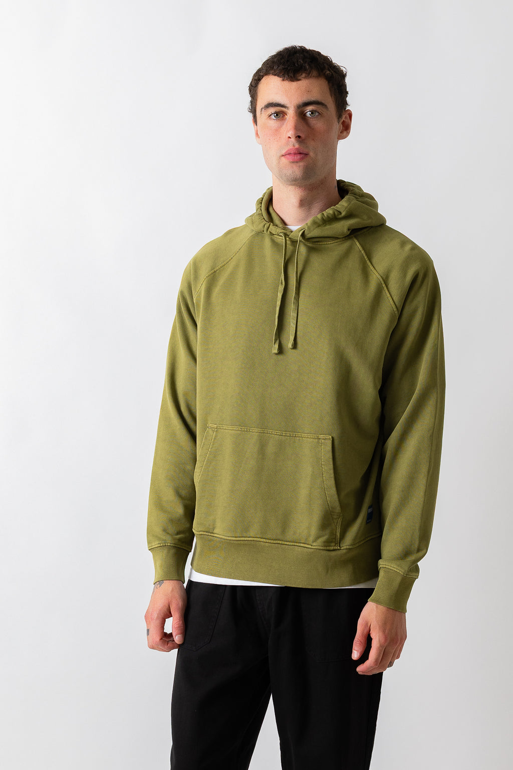 A man wearing an olive green pullover hoodie with a drawstring hood and kangaroo pocket, paired with black pants, standing against a light gray studio background.