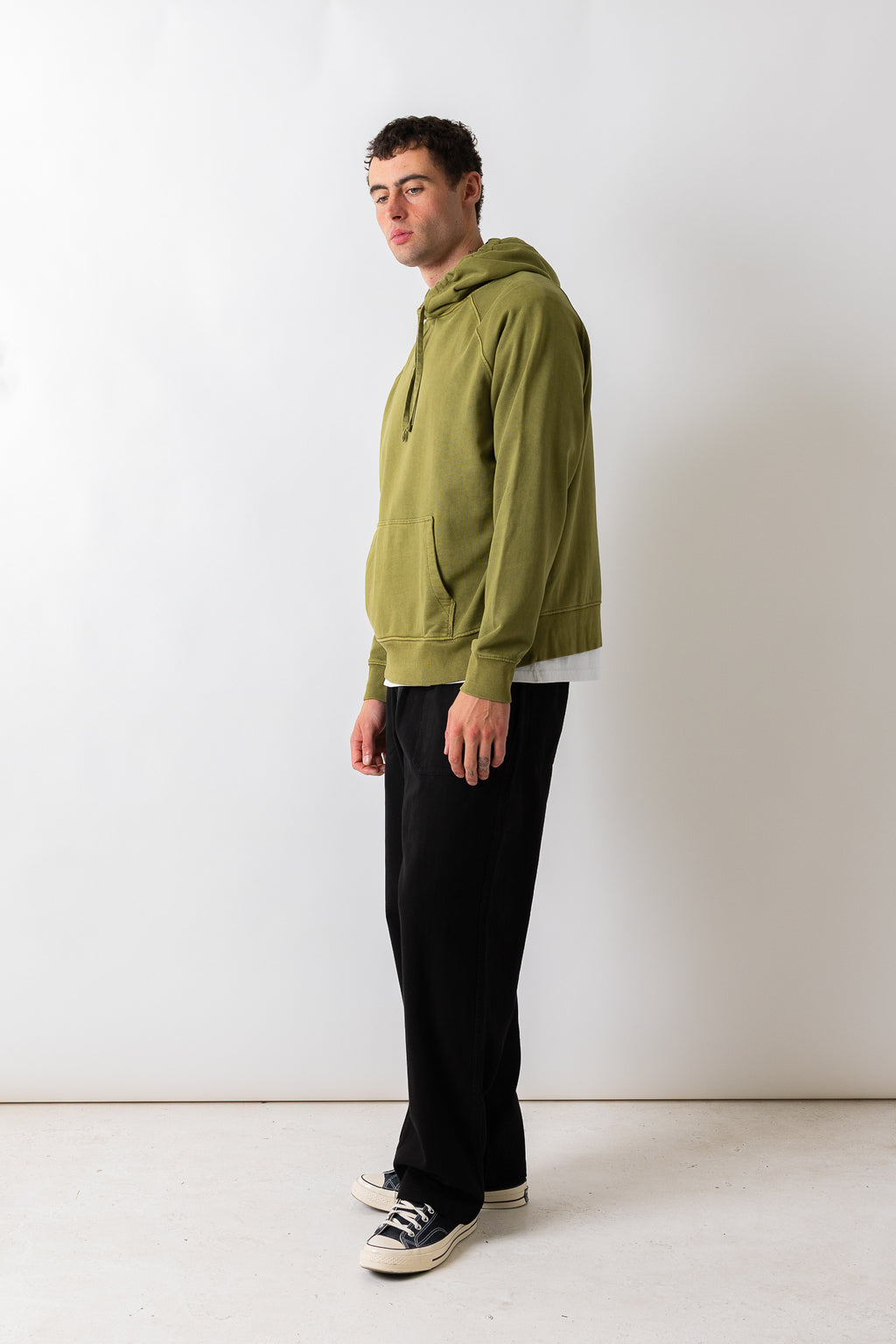 A man wearing an olive green pullover hoodie, black wide-leg trousers, and black-and-white sneakers, standing in profile against a plain white studio wall.
