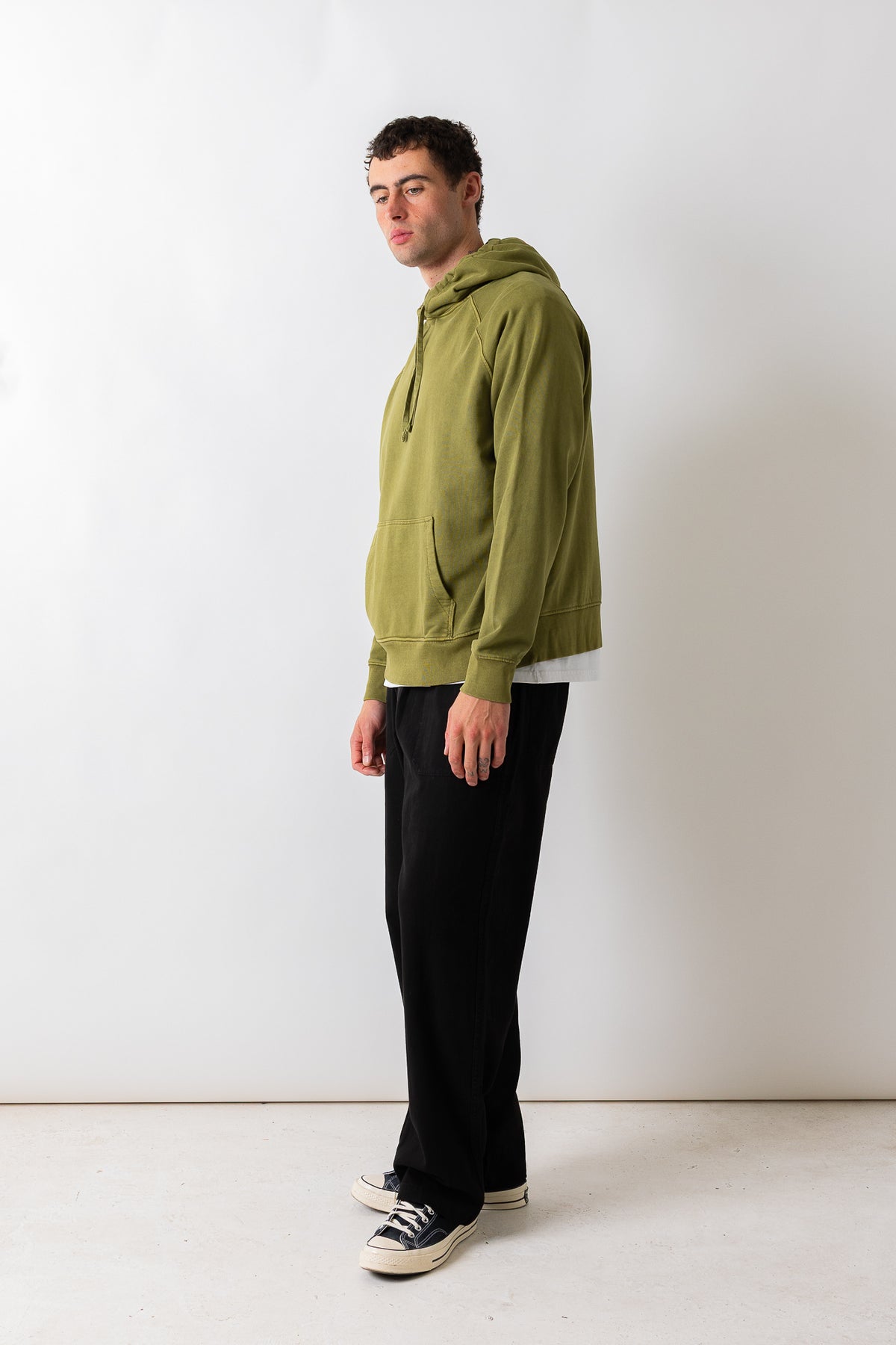 A man wearing an olive green pullover hoodie, black wide-leg trousers, and black-and-white sneakers, standing in profile against a plain white studio wall.