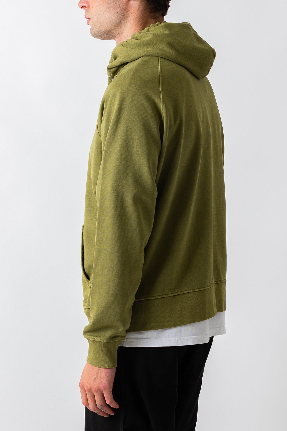 A man shown from the side profile wearing an olive green pullover hoodie over a white t-shirt and black pants, photographed against a plain light gray studio background.