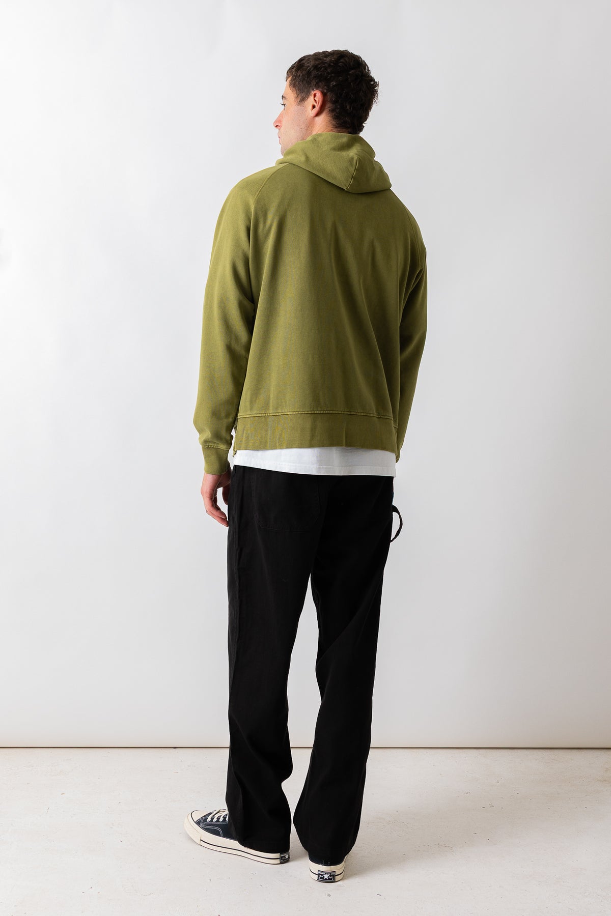 A man is shown from the back wearing an olive green pullover hoodie, black sweatpants, and black-and-white sneakers against a plain white studio background.