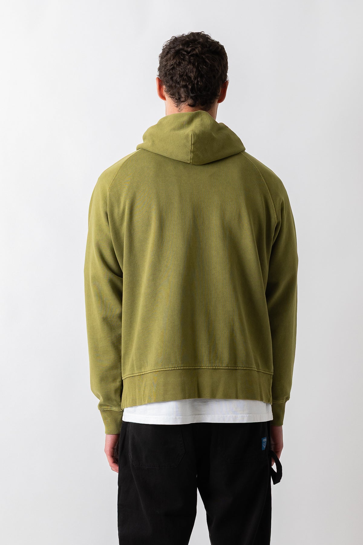 A man viewed from behind wearing an olive green pullover hoodie over a white t-shirt and black pants, standing against a plain light gray background.
