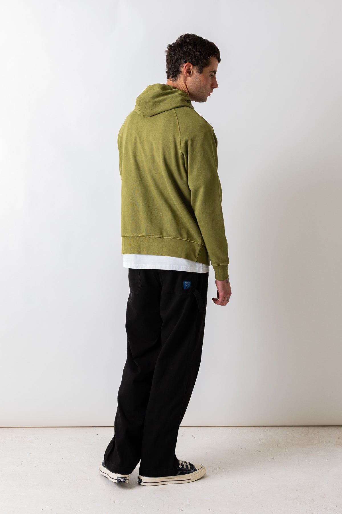 A man wearing an olive green hooded sweatshirt, black sweatpants, and black-and-white sneakers, standing in profile against a plain white studio wall.