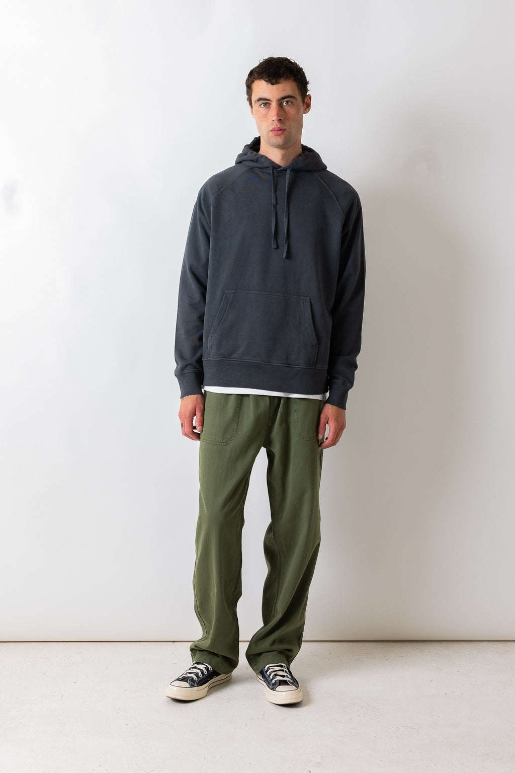 A man stands against a plain white studio wall wearing a dark charcoal gray pullover hoodie, olive green pants, and black-and-white sneakers.