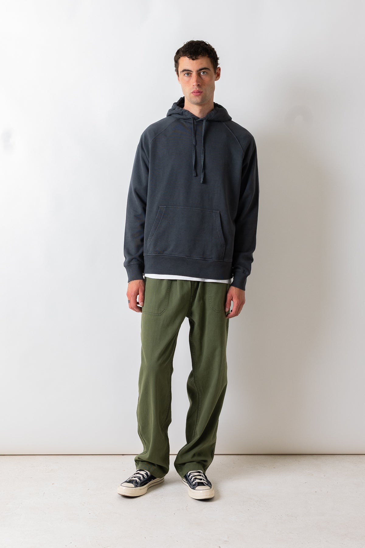 A man stands against a plain white studio wall wearing a dark charcoal gray pullover hoodie, olive green pants, and black-and-white sneakers.