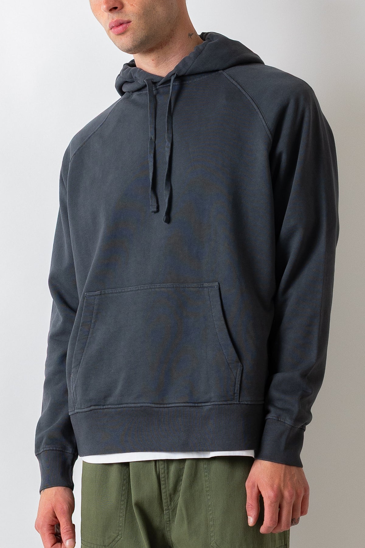 A man wearing a dark charcoal gray hooded sweatshirt with a subtle tonal graphic pattern, paired with olive green pants, shown from the chin down against a light gray background.