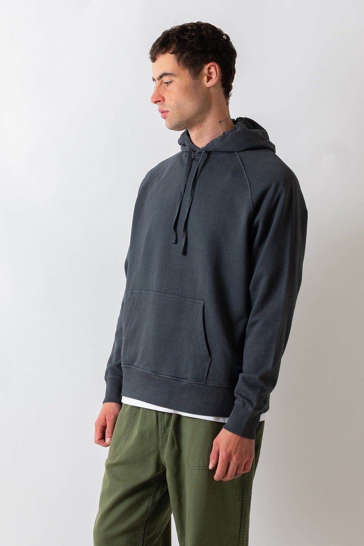 A man wearing a dark charcoal gray hooded sweatshirt and olive green pants, standing in profile against a light gray studio background.