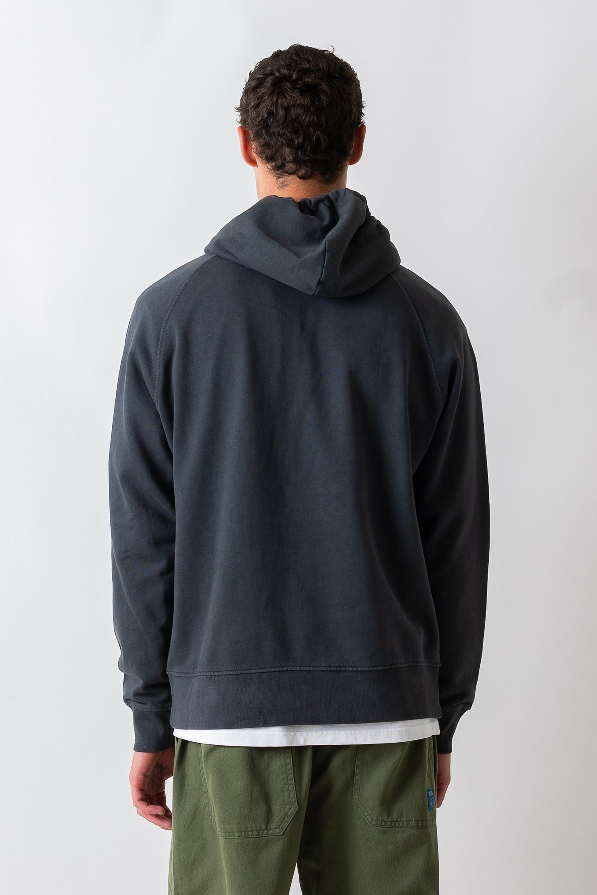 A man viewed from behind wearing a dark charcoal gray hooded sweatshirt, olive green pants, and a white t-shirt layered underneath, set against a plain light gray studio background.