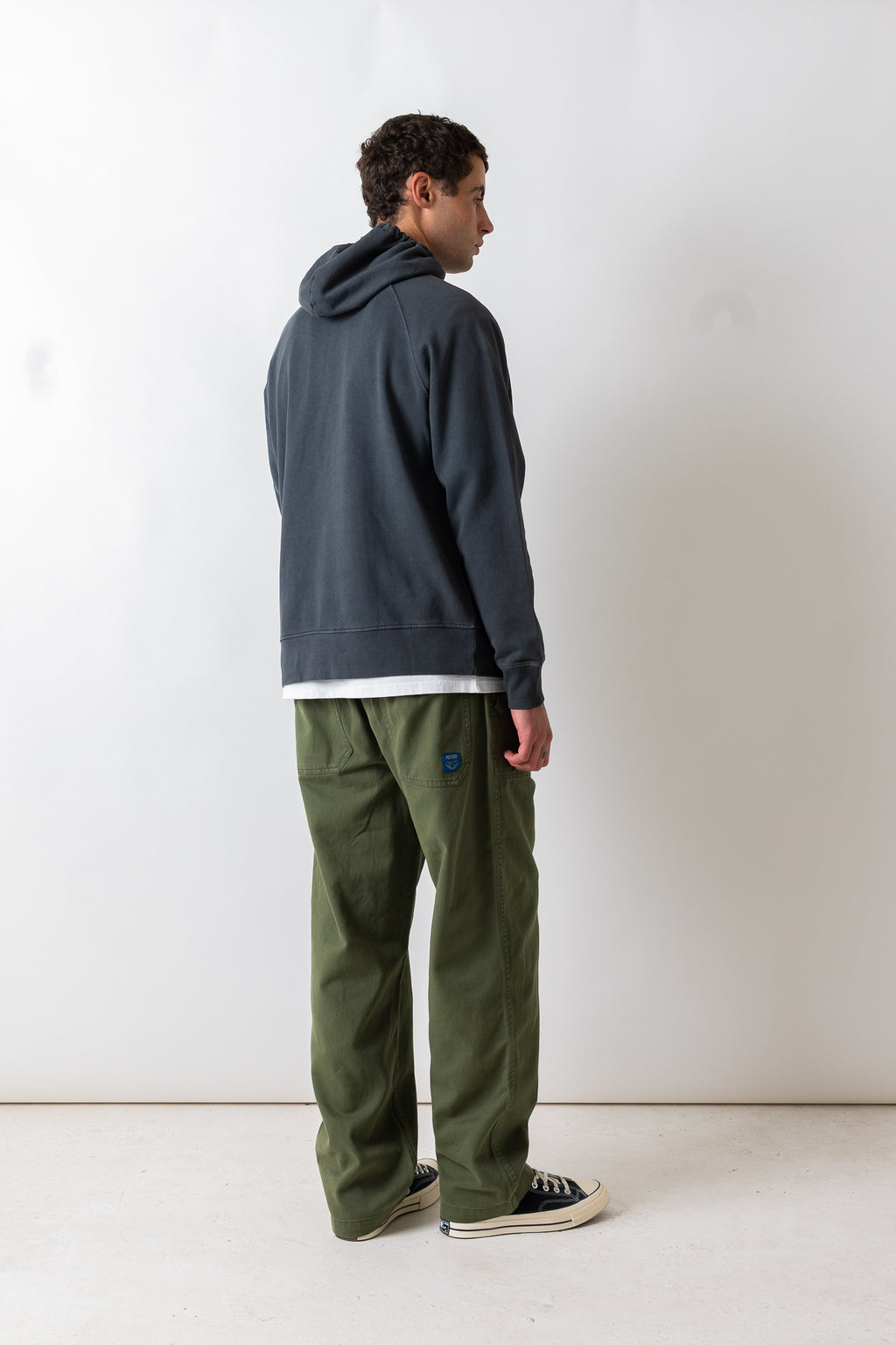 A man standing in profile wearing a dark slate blue hoodie, olive green pants, and black canvas sneakers against a plain white studio background.
