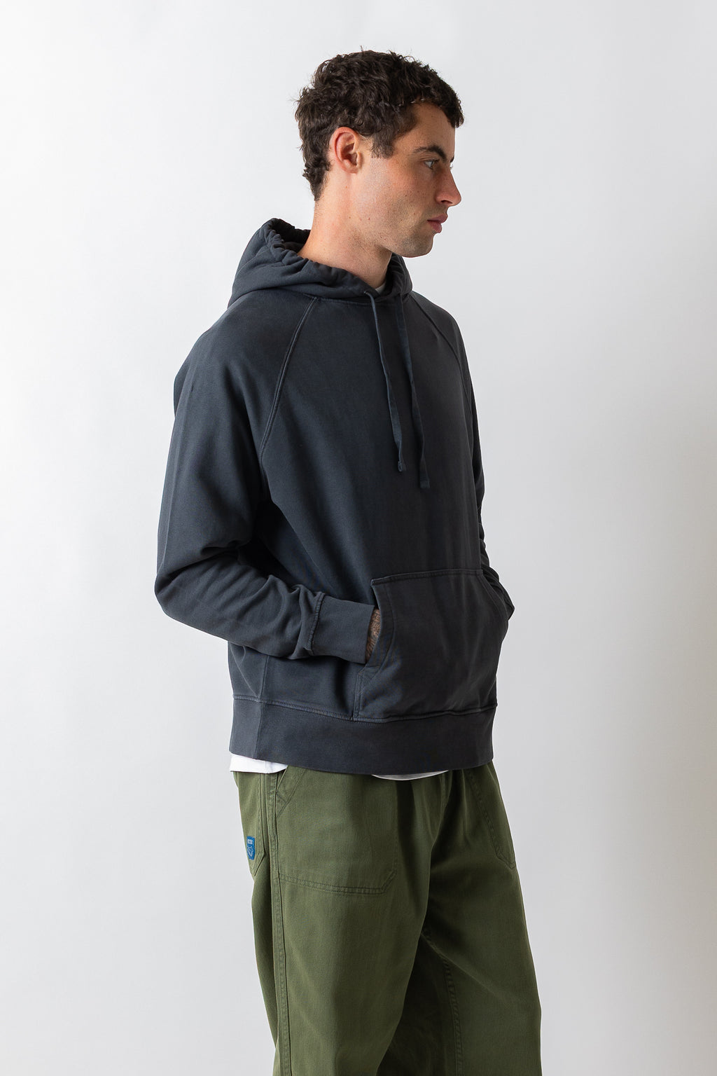 A man wearing a dark navy pullover hoodie with a kangaroo pocket and olive green pants, standing in profile against a light gray studio background.