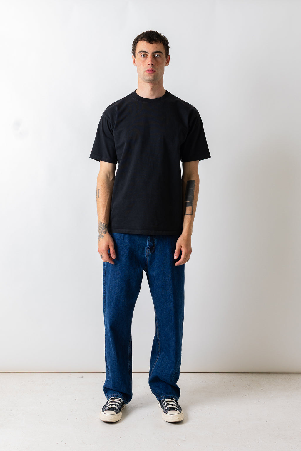 A man wearing a plain black short-sleeve t-shirt, blue jeans, and black-and-white sneakers, standing against a plain off-white studio wall.