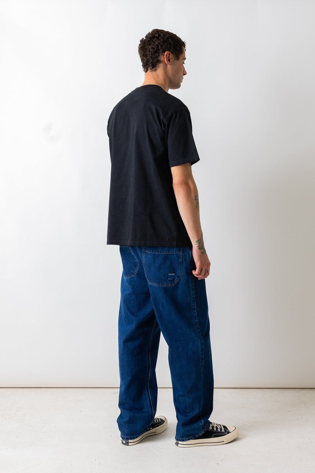 A man shown from the back wearing a black short-sleeve t-shirt, blue denim jeans, and black canvas sneakers, standing against a plain white studio wall.