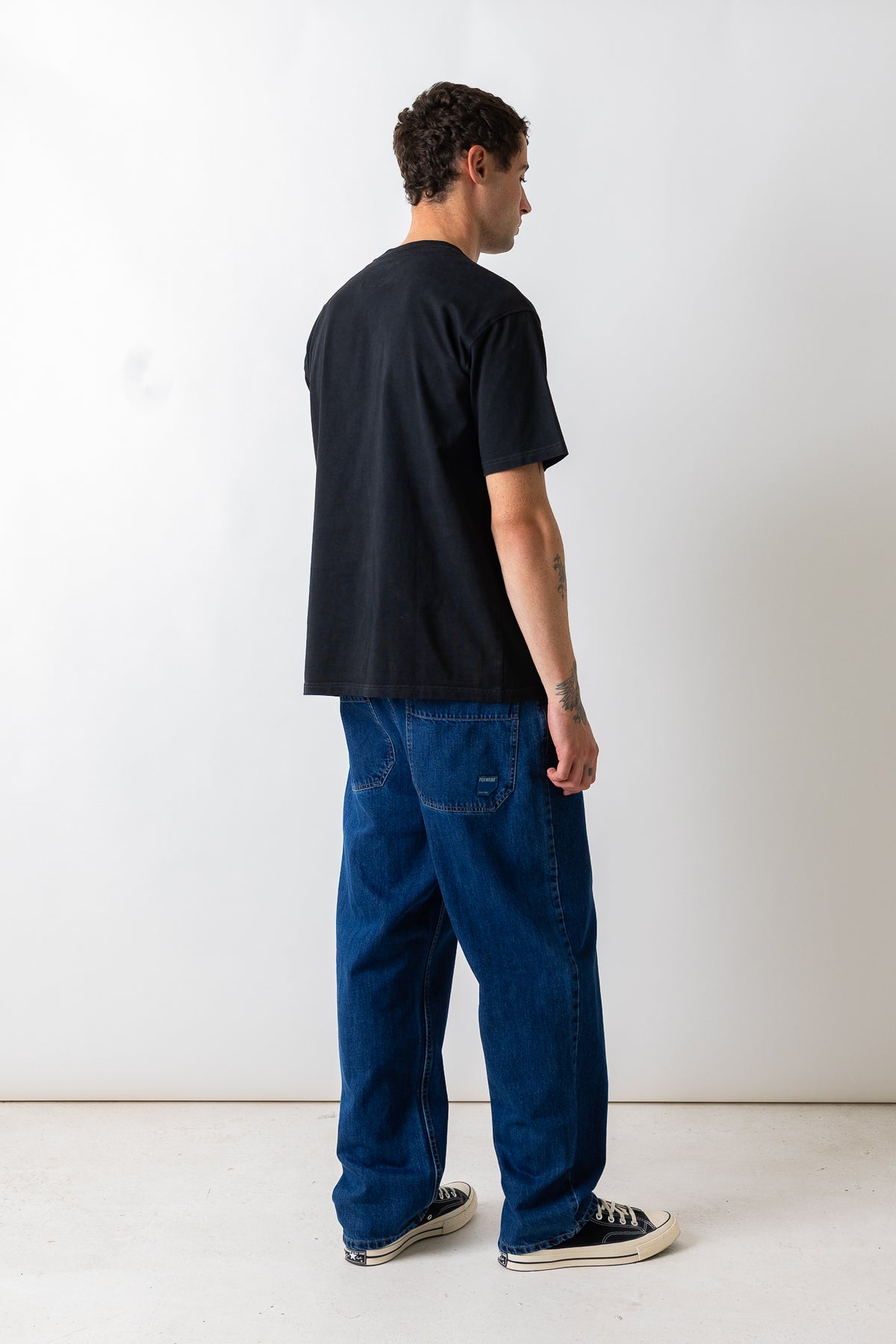 A man shown from the back wearing a black short-sleeve t-shirt, blue denim jeans, and black canvas sneakers, standing against a plain white studio wall.