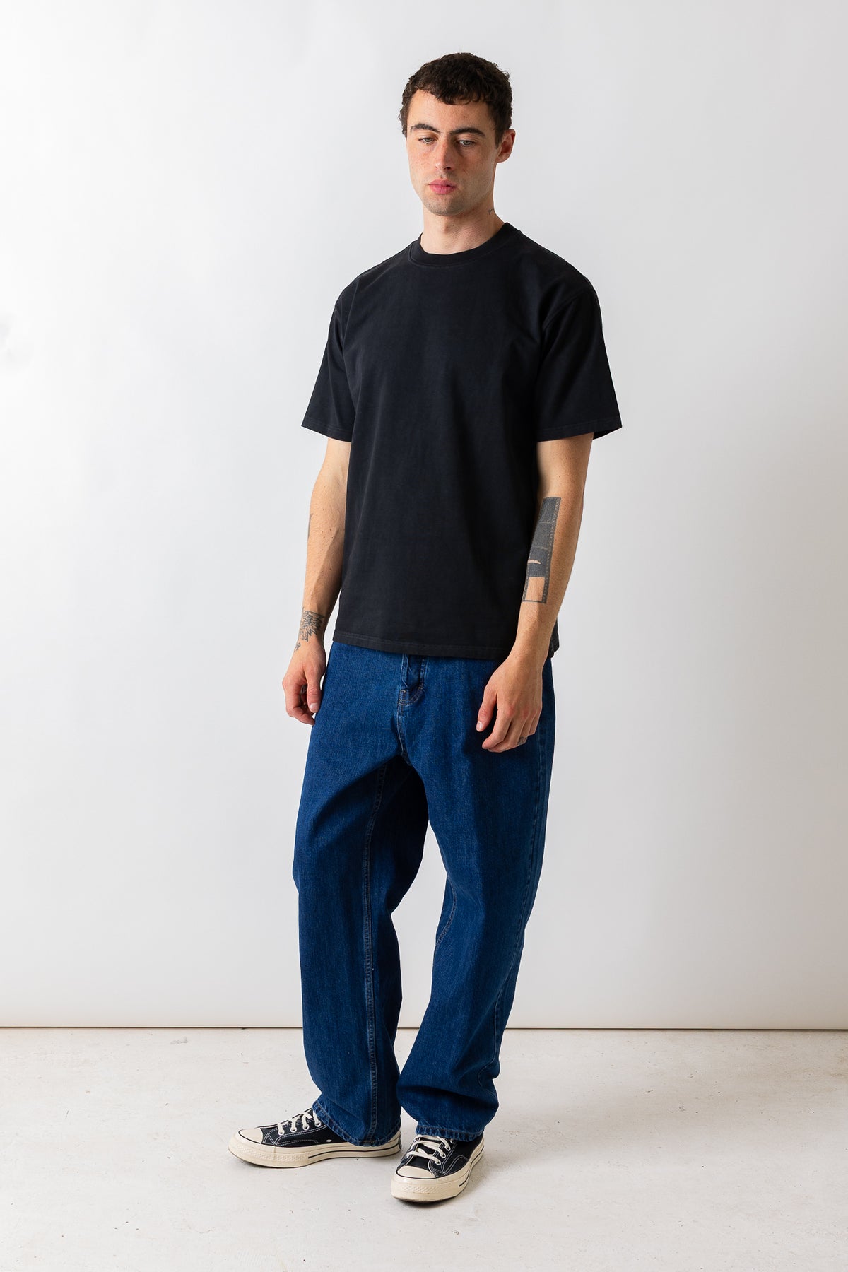 A man modeling a black short-sleeve crewneck t-shirt and loose-fit dark blue jeans, paired with black and white canvas sneakers, standing against a plain white studio background.