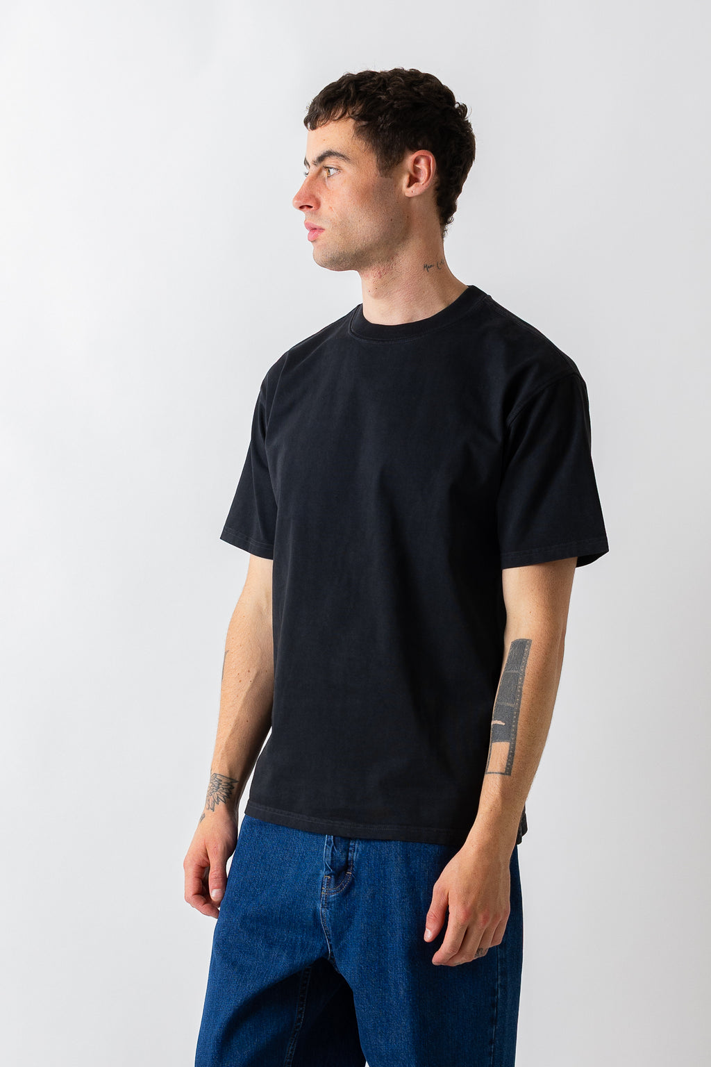A man wearing a plain black short-sleeve crewneck t-shirt and blue jeans, standing against a light gray studio background.