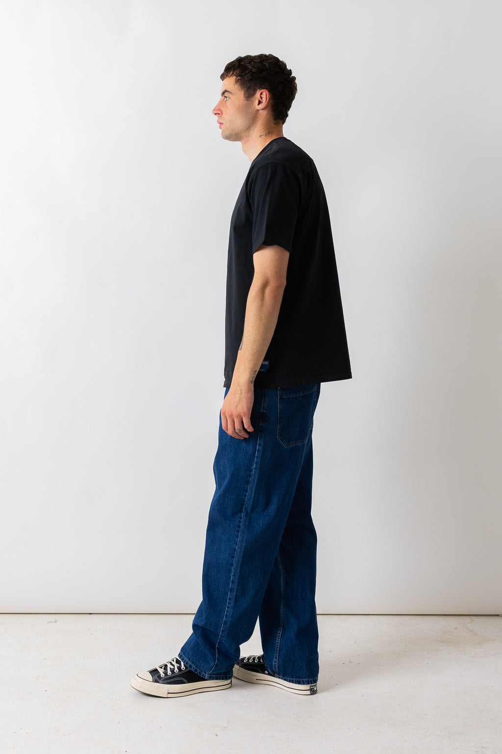A man standing in profile wearing a black short-sleeve t-shirt, loose blue jeans, and black canvas sneakers against a plain white studio background.