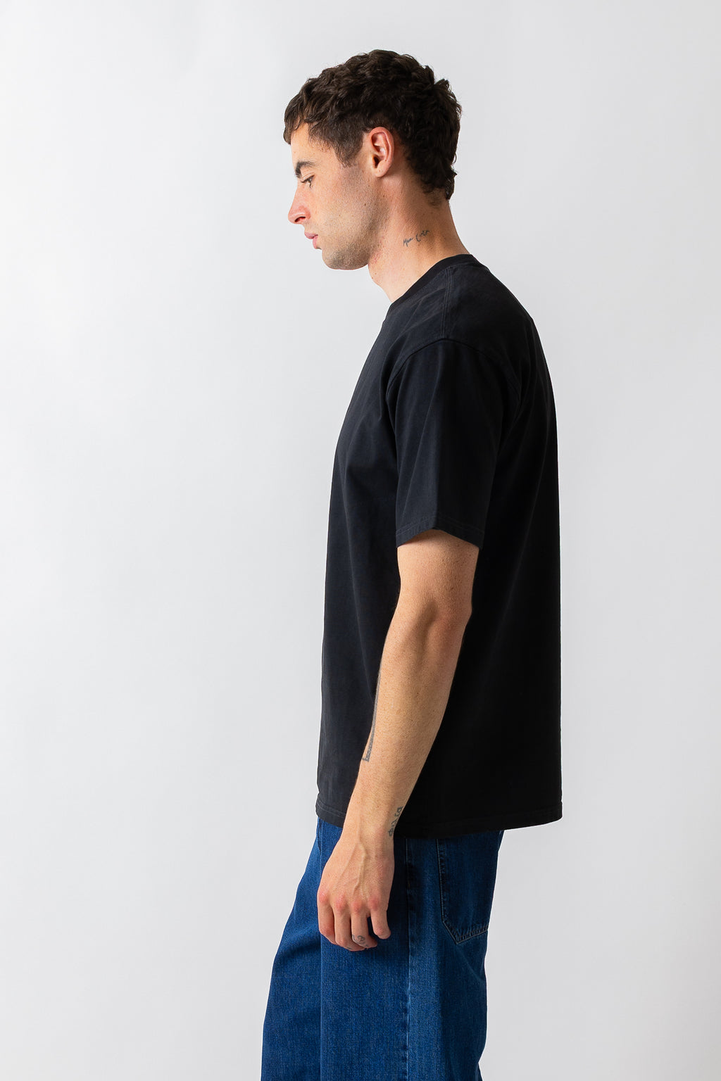 A man in profile wearing a plain black short-sleeve t-shirt and blue jeans, standing against a light gray studio background.