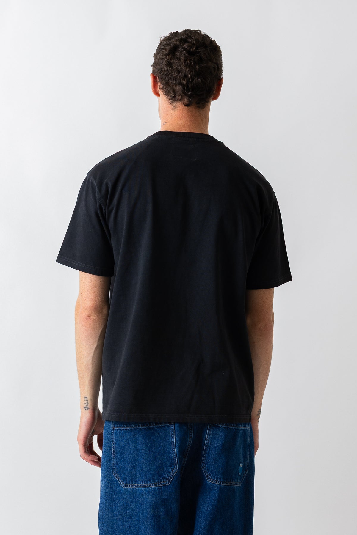 A man viewed from behind wearing a plain black short-sleeve t-shirt and blue jeans against a light gray studio background.