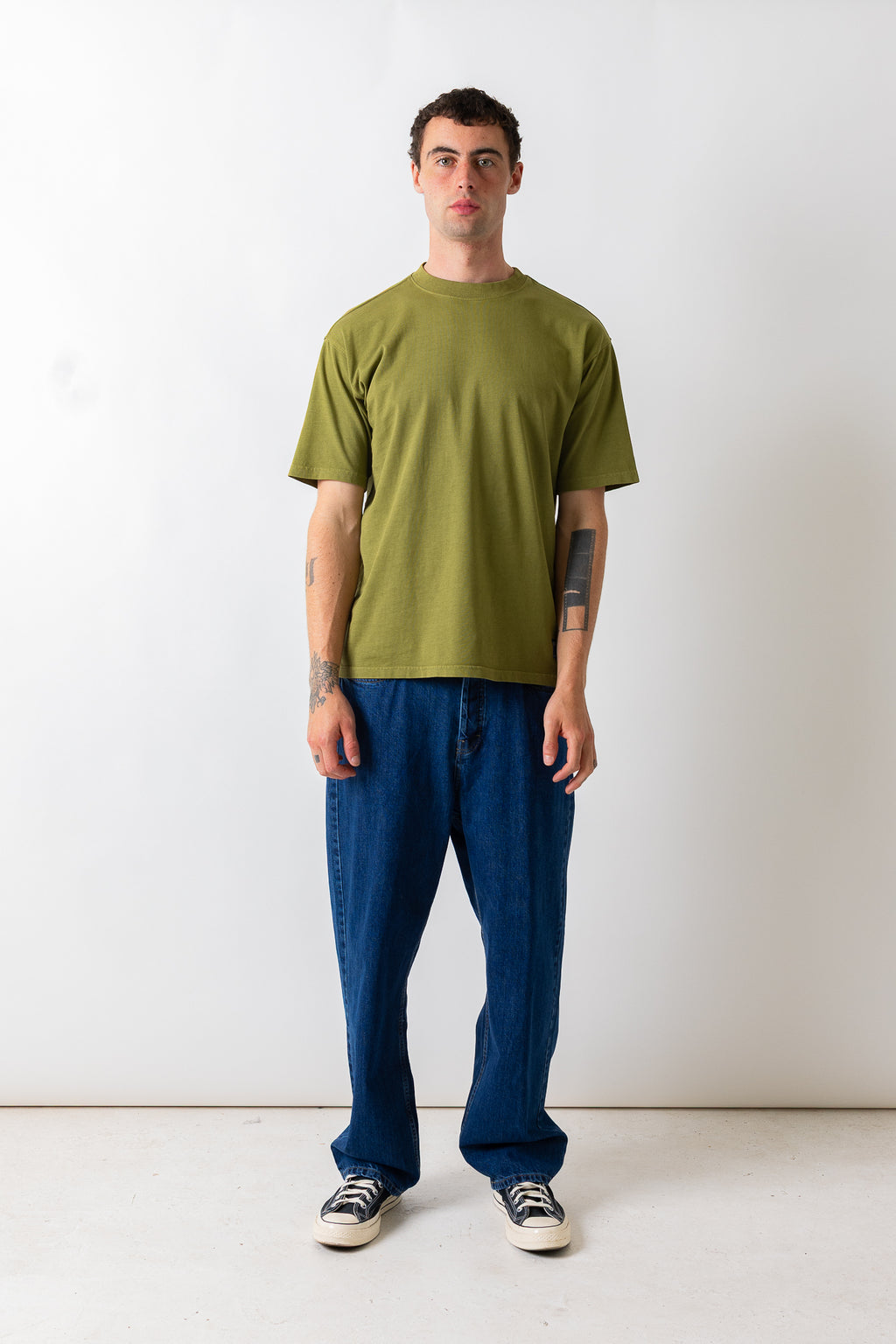 A man stands against a plain white studio wall wearing an olive green short-sleeve t-shirt, loose blue jeans, and black-and-white sneakers.
