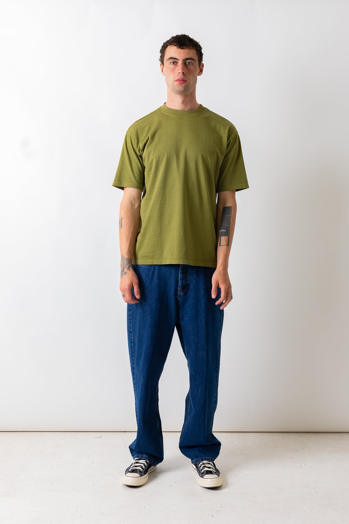 A man stands against a plain white studio wall wearing an olive green short-sleeve t-shirt, loose blue jeans, and black-and-white sneakers.