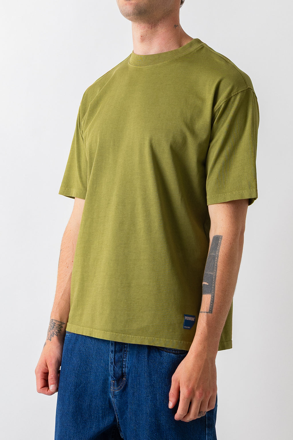 A man wearing an olive green short-sleeve crewneck t-shirt and blue jeans, shown from the neck down against a light gray background.