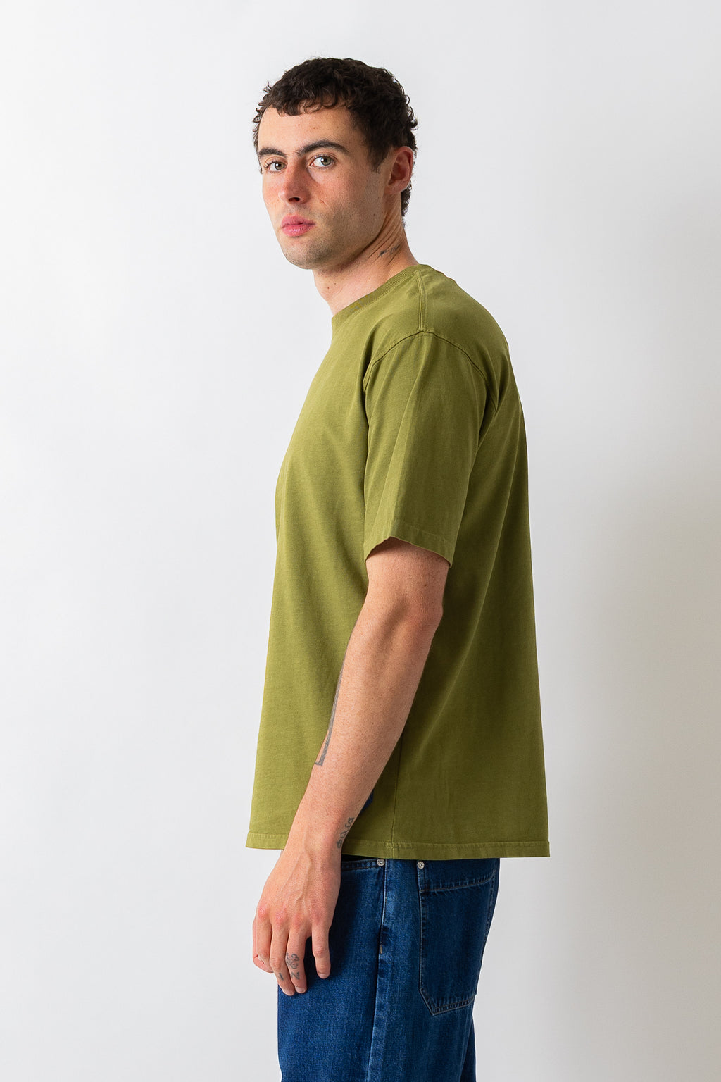 A man wearing a muted olive green short-sleeve t-shirt and blue jeans, shown in profile against a plain light gray studio background.