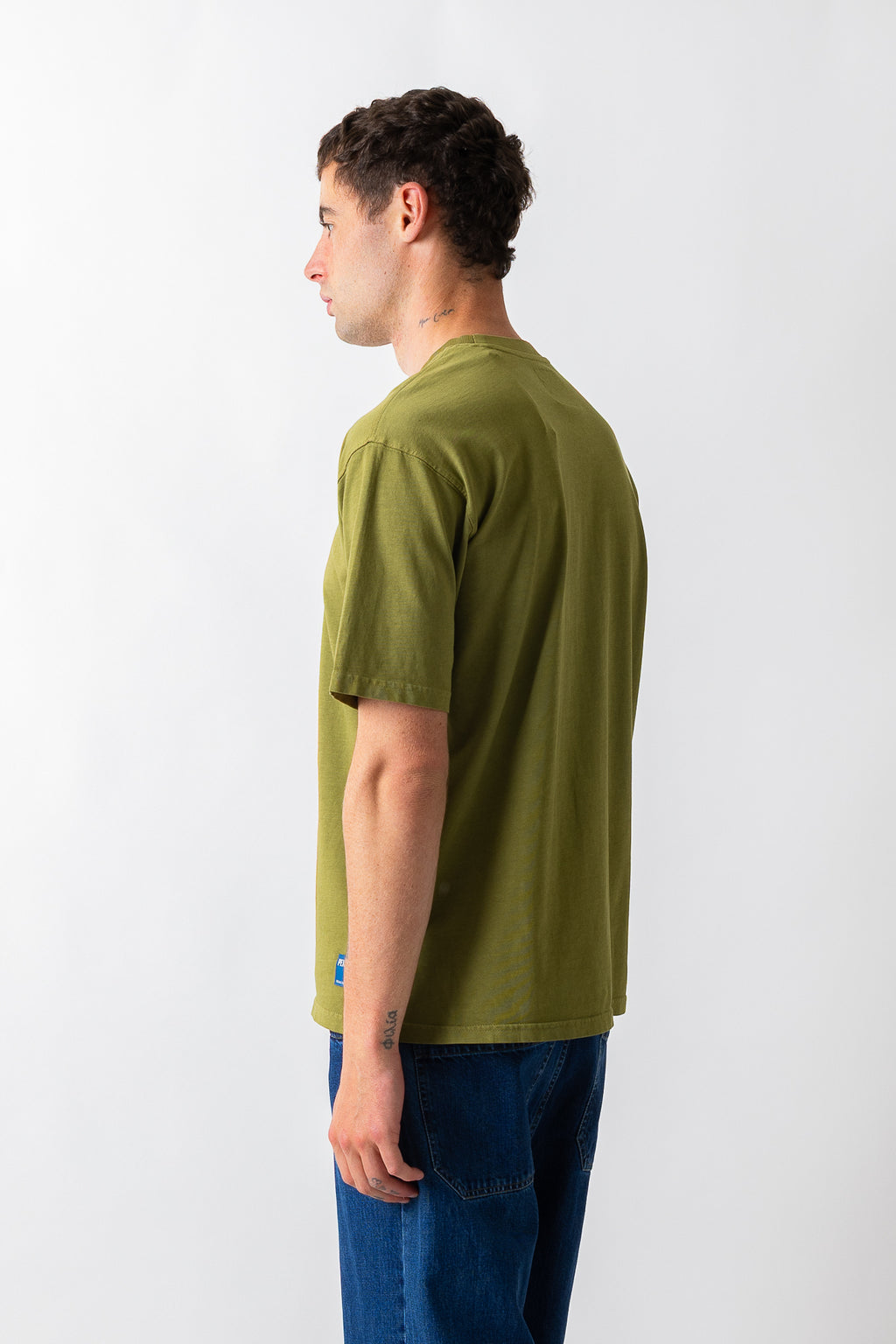 A man shown from the side profile wearing an olive green short-sleeve t-shirt and blue jeans, standing against a plain white studio background.