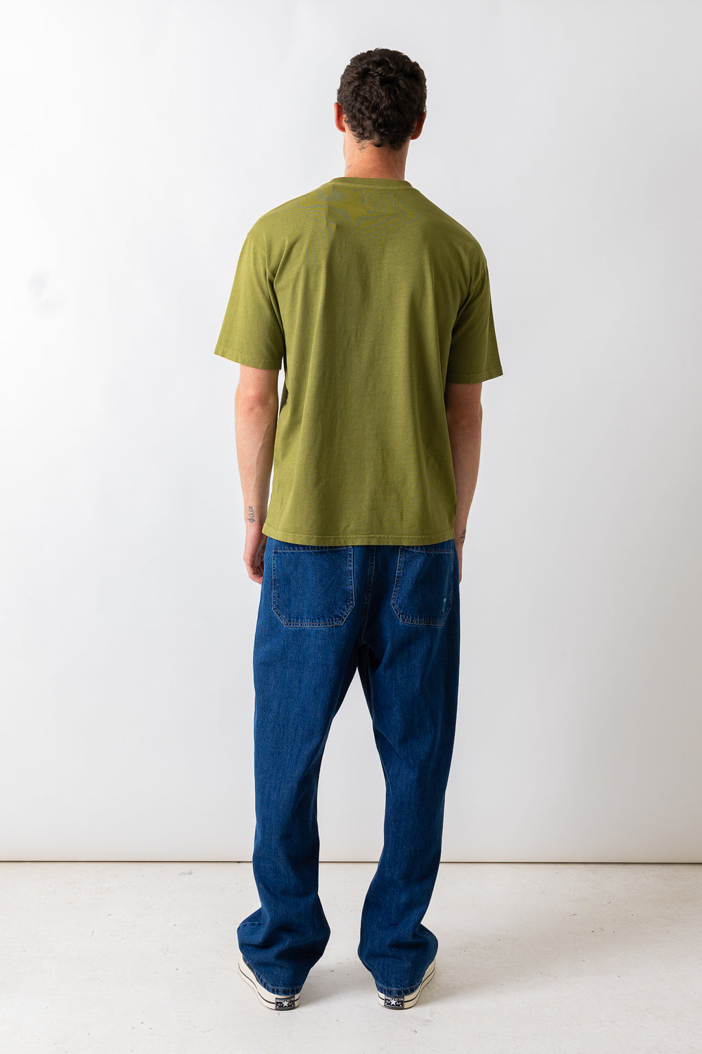 A man viewed from behind wearing an olive green short-sleeve t-shirt and blue jeans, standing against a plain white studio background.