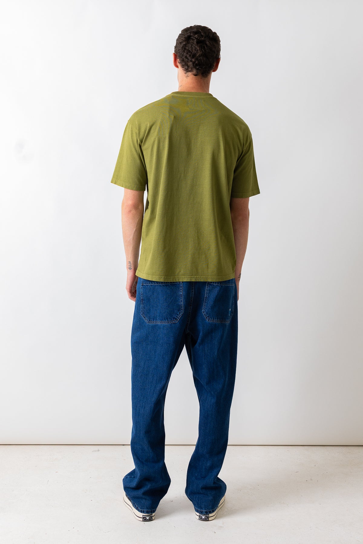 A man viewed from behind wearing an olive green short-sleeve t-shirt and blue jeans, standing against a plain white studio background.