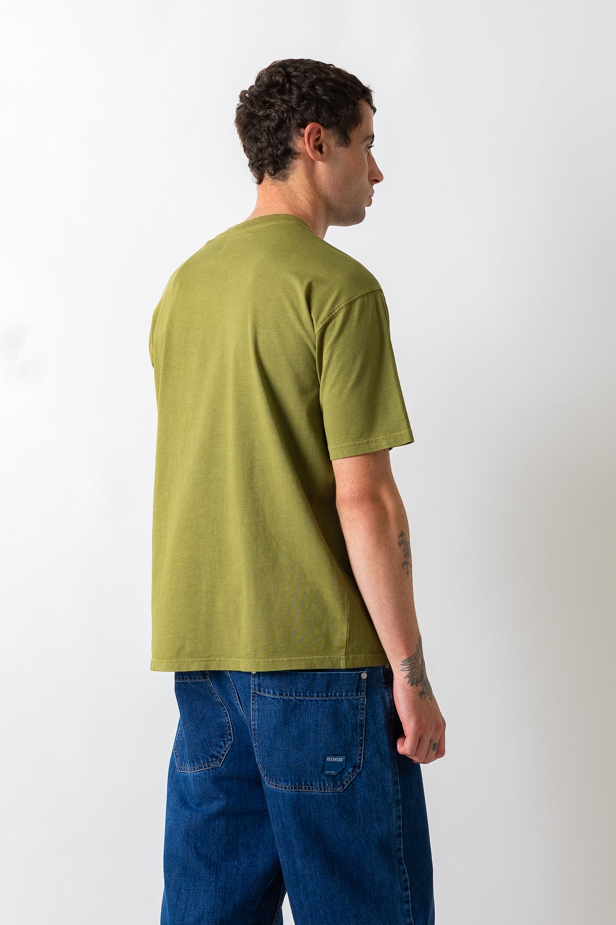 A man wearing a muted olive green short-sleeve t-shirt and blue jeans, shown from a side-back angle against a plain white studio background.