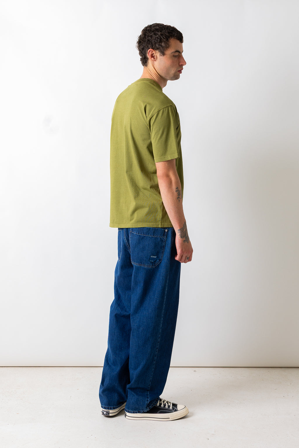 A man standing in profile wearing an olive green short-sleeve t-shirt, loose-fit dark blue jeans, and black canvas sneakers against a plain white studio background.