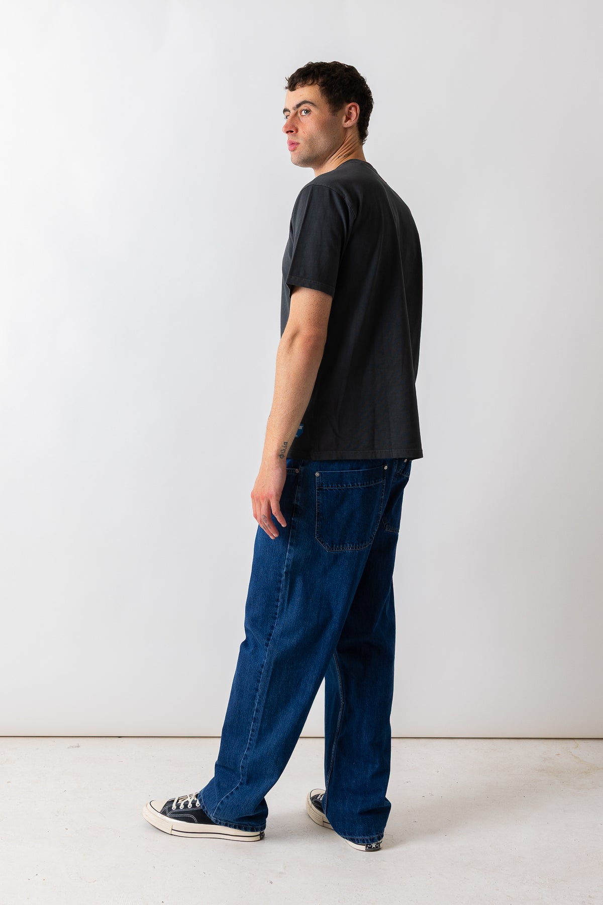 A man standing in profile wearing a faded black t-shirt, dark blue denim jeans, and black canvas sneakers against a plain white studio background.