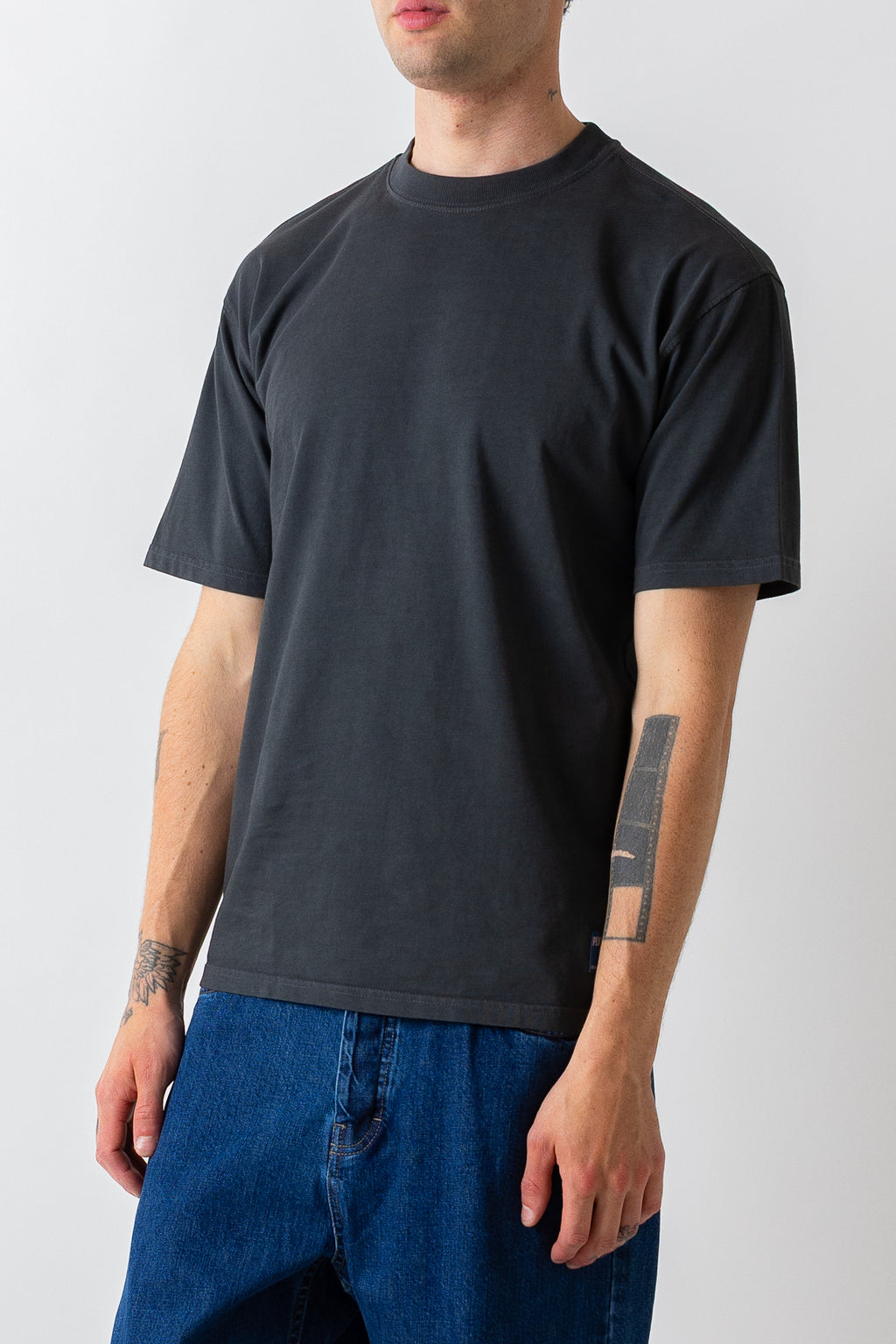 A man wearing a dark charcoal gray crewneck t-shirt and blue jeans, shown from the waist up against a light gray background.