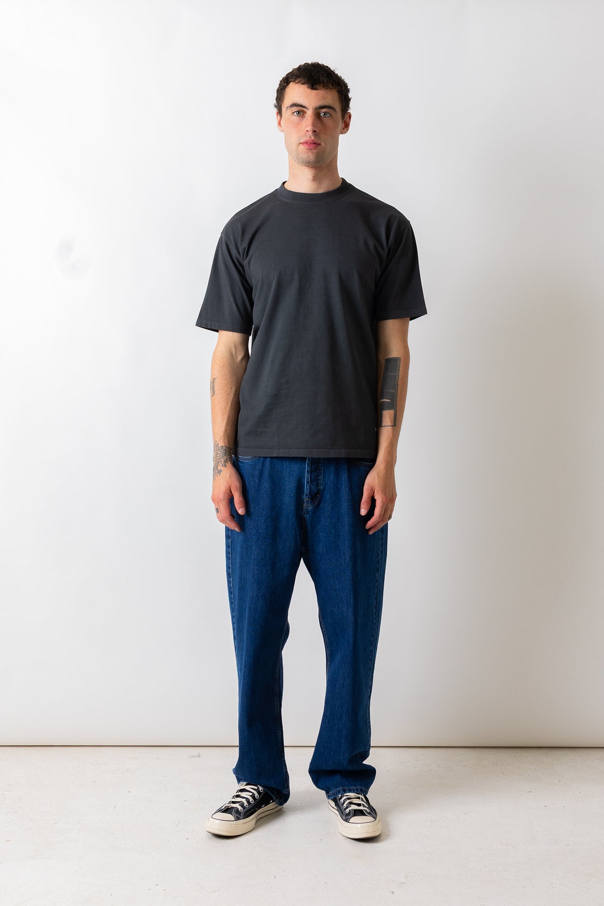 A man stands against a plain white studio wall wearing a dark charcoal gray crewneck t-shirt, loose blue jeans, and black-and-white sneakers.