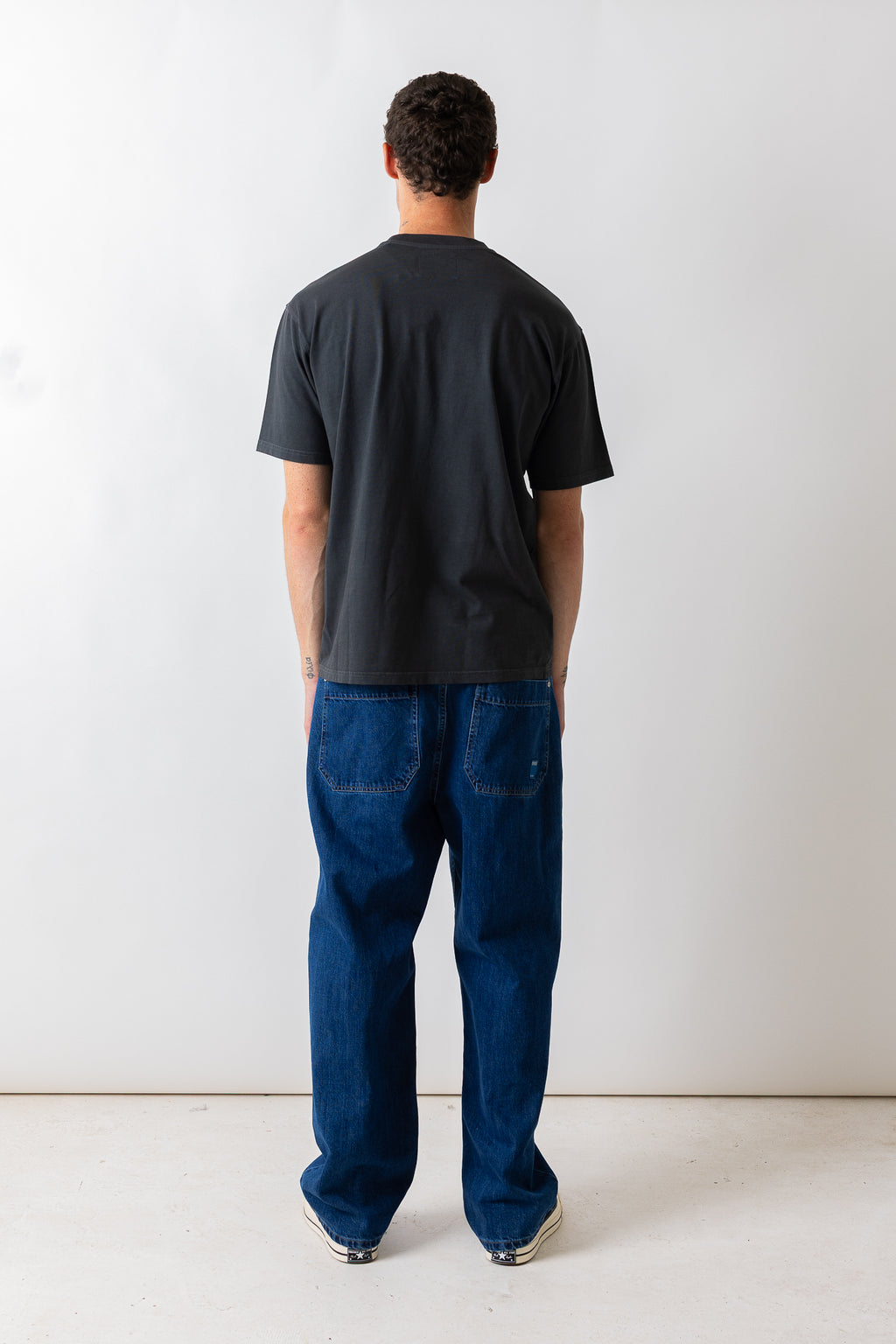 A man viewed from behind wearing a dark charcoal gray t-shirt and blue jeans, standing against a plain white studio background.
